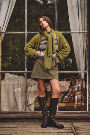 A woman stands by large window panes, wearing the RINA ALPACA SWEATER by Velvet by Graham & Spencer in green, paired with a matching scarf, olive skirt, and black boots. Blankets and curtains are visible inside.