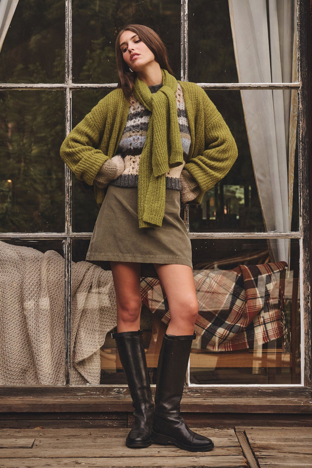 A woman stands on a wooden deck wearing a green sweater, striped top, olive skirt, black knee-high boots, and the LUCA ALPACA SCARF from Velvet by Graham & Spencer. A plaid blanket is visible through a window behind her.