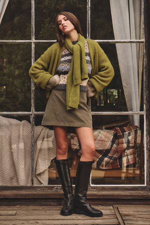 A woman stands on a wooden deck wearing a green sweater, striped top, olive skirt, black knee-high boots, and the LUCA ALPACA SCARF from Velvet by Graham & Spencer. A plaid blanket is visible through a window behind her.