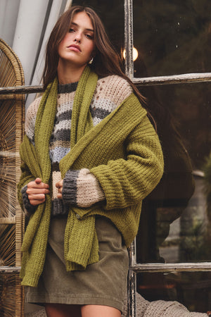 A woman stands by a window wearing a chunky green cardigan, striped knit sweater, olive skirt, and the LUCA ALPACA SCARF by Velvet by Graham & Spencer—a perfect winter accessory.