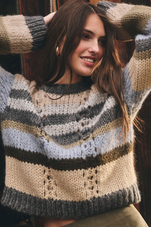 A woman smiles with her arms raised behind her head, wearing the RINA ALPACA SWEATER by Velvet by Graham & Spencer—a boxy fit, chunky knit striped sweater in beige, gray, and blue tones.