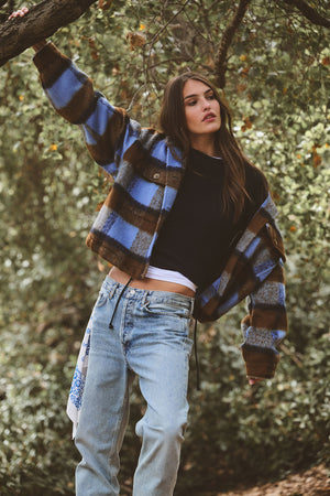 A woman stands outdoors under a tree, wearing the SAVANNAH SWEATER by Velvet by Graham & Spencer—a wool blend crewneck—layered over a white shirt with a blue and brown plaid jacket and light blue jeans.