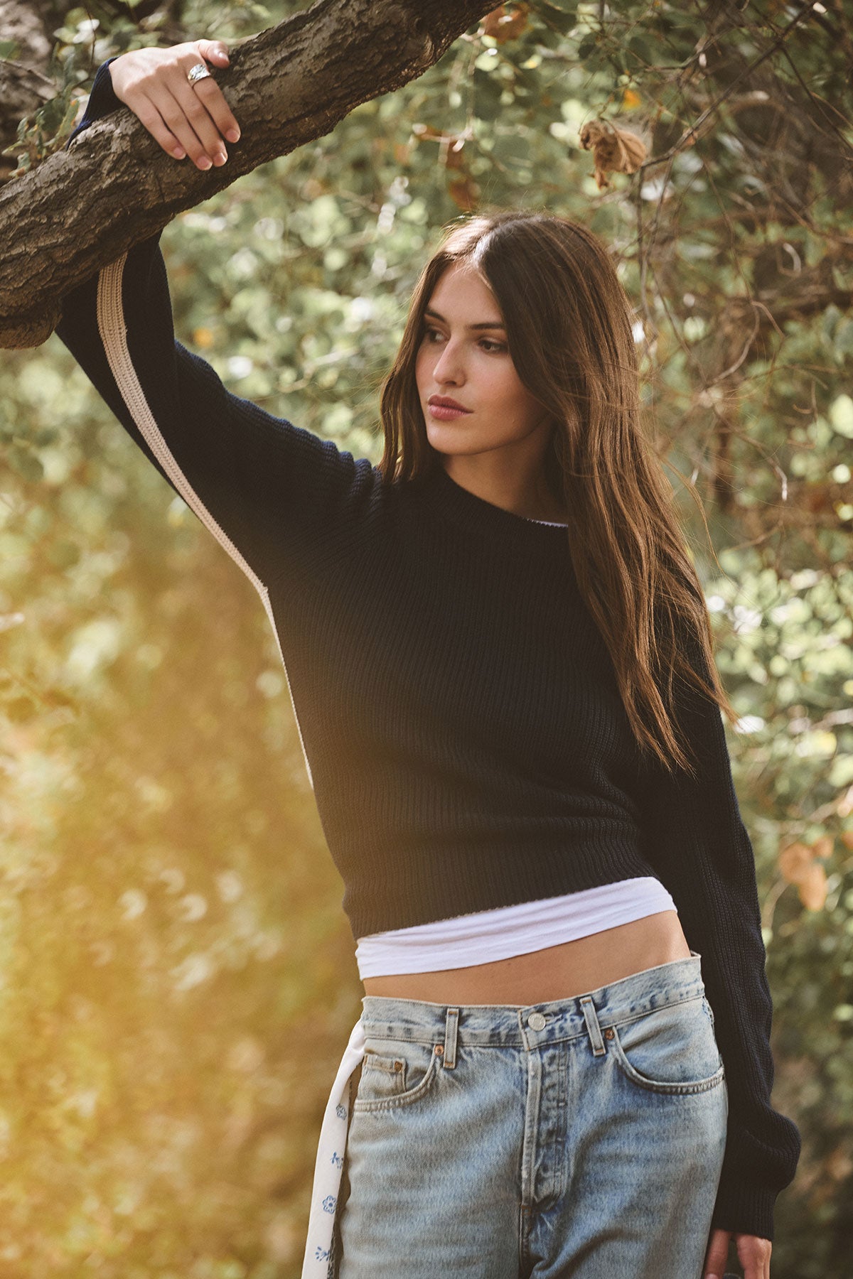 A woman with long brown hair wears the Velvet by Graham & Spencer SAVANNAH SWEATER, paired with a white shirt and jeans, as she leans against a tree outdoors surrounded by greenery.-39839775457473