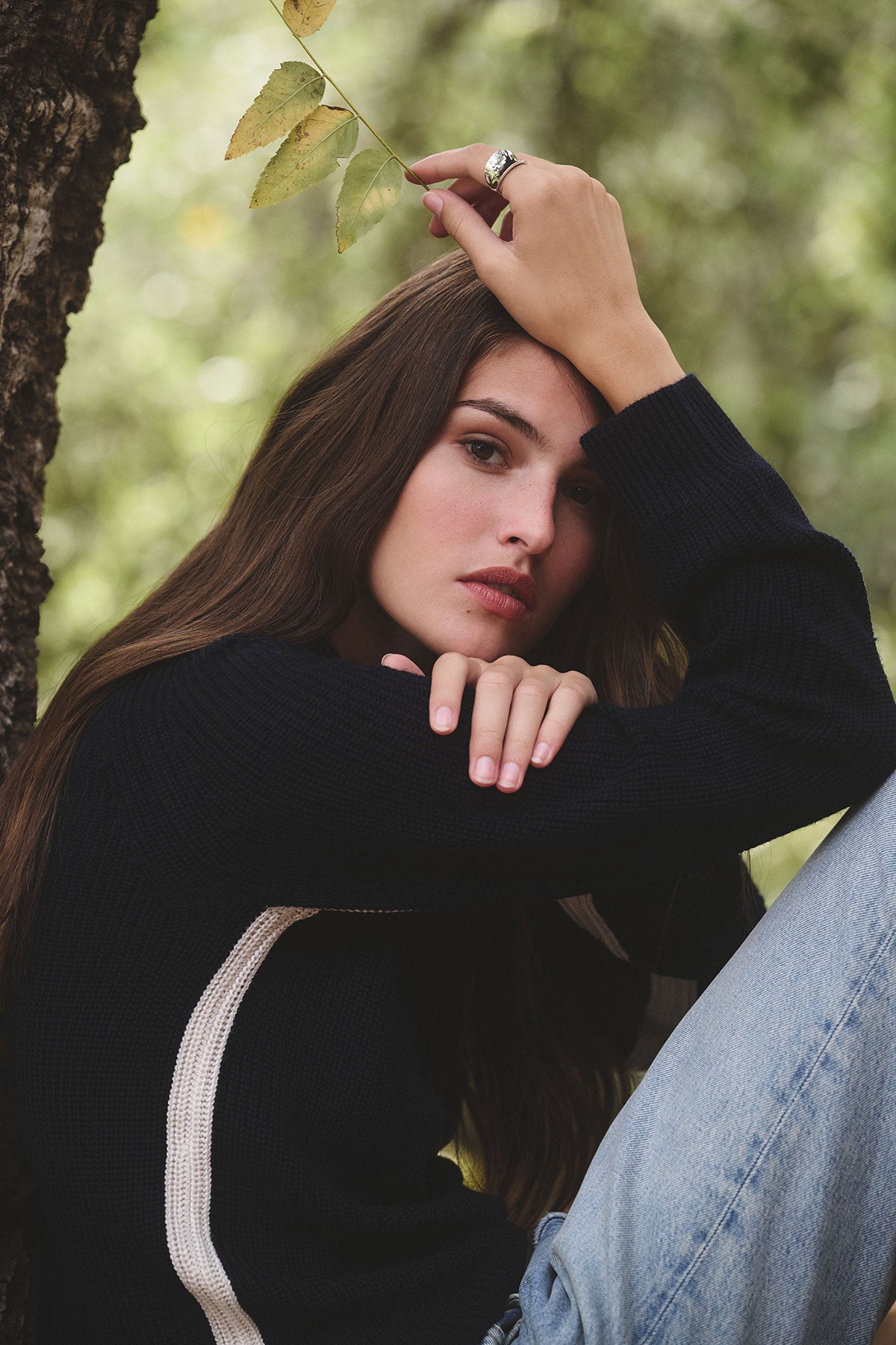 A woman with long brown hair, wearing the SAVANNAH SWEATER by Velvet by Graham & Spencer and jeans, sits by a tree, holding a small branch above her head.-39839771885761