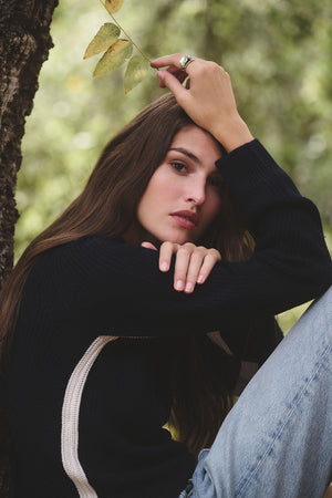 A woman with long brown hair, wearing the SAVANNAH SWEATER by Velvet by Graham & Spencer and jeans, sits by a tree, holding a small branch above her head.
