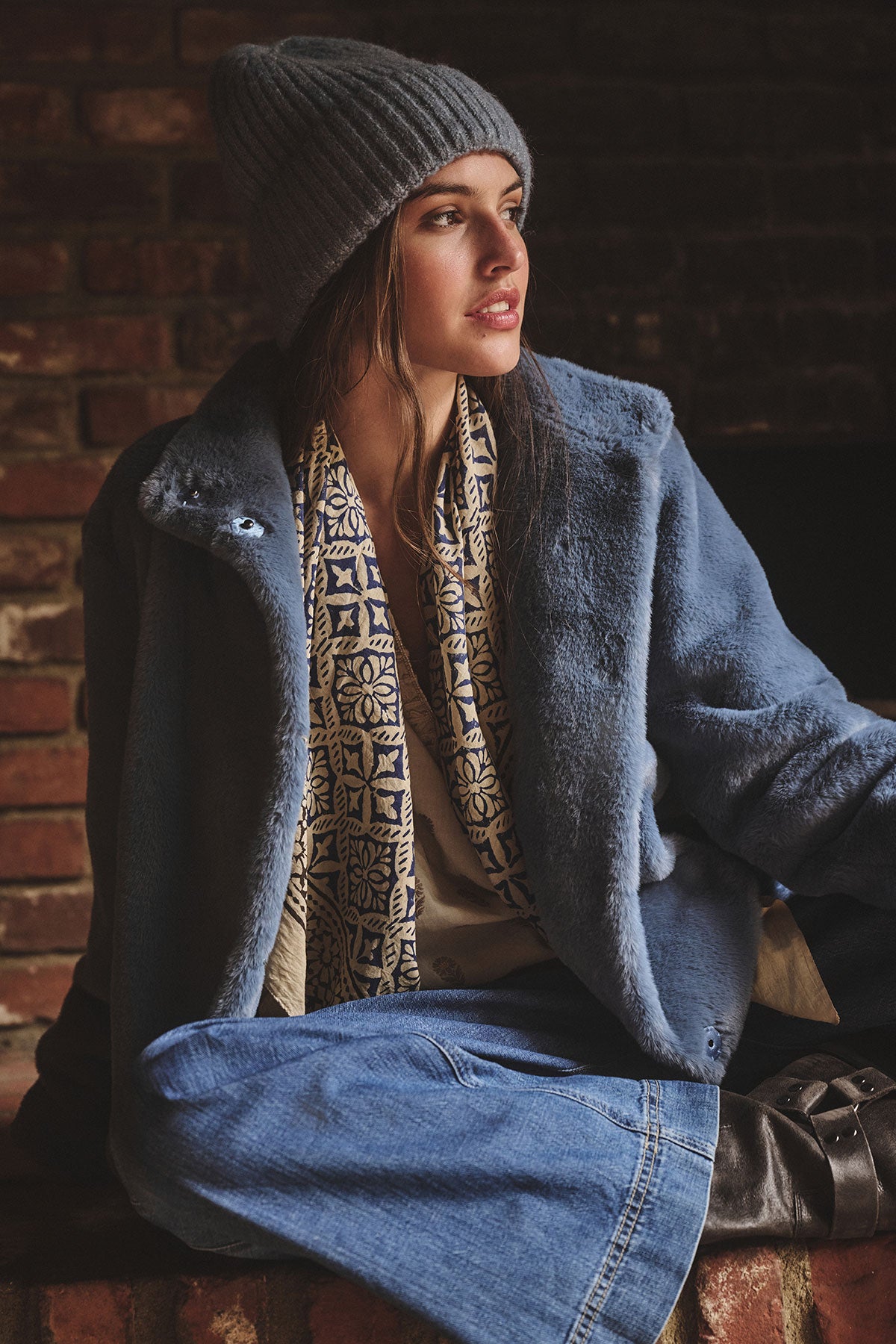 A woman wears the KEY BEANIE by Velvet by Graham & Spencer, a blue faux fur coat, patterned scarf, and denim skirt as she sits indoors against a brick wall looking to the side.-39850403954881