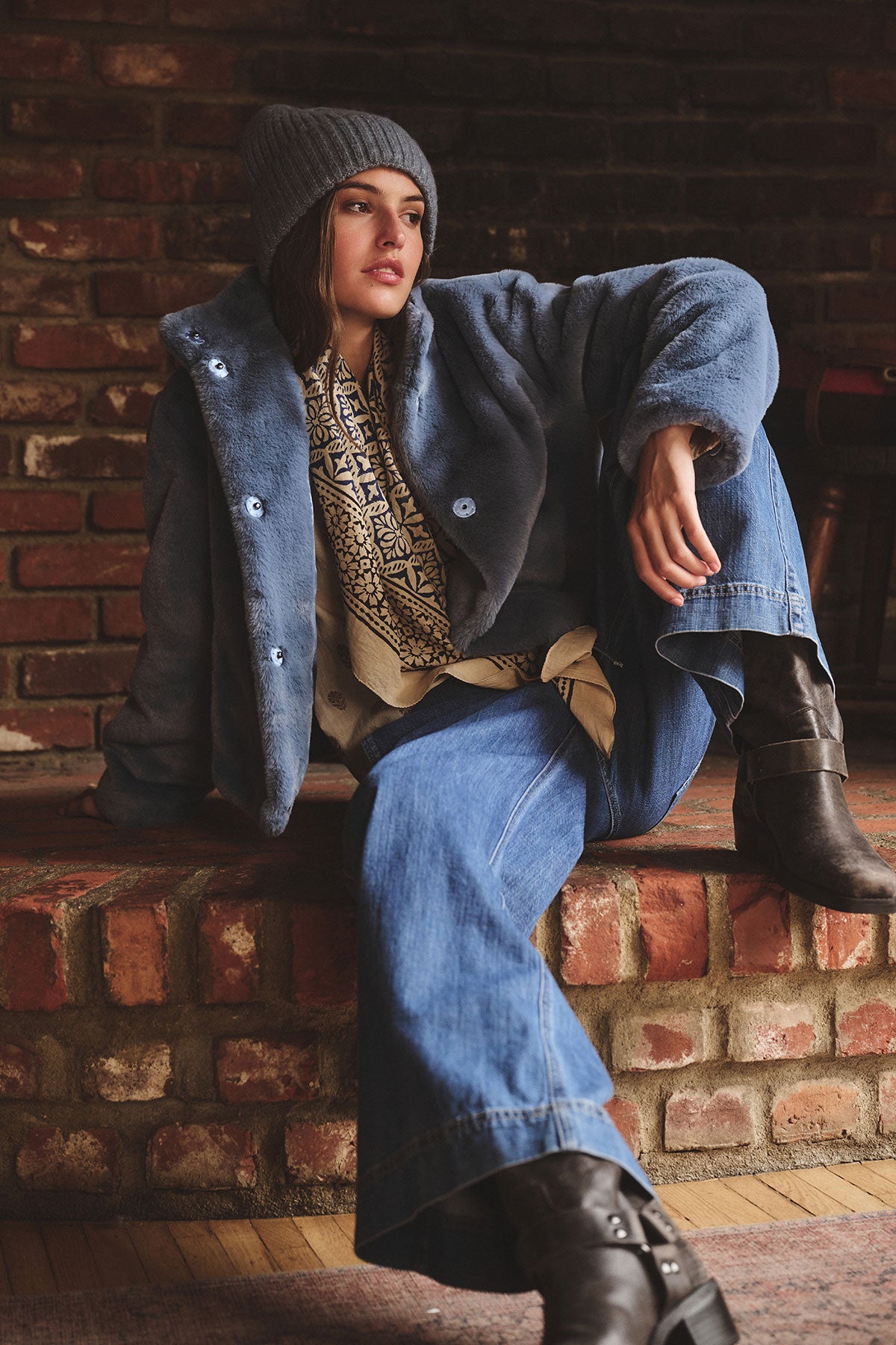   A person in a SHEENA LUXE FAUX FUR JACKET by Velvet by Graham & Spencer, wide-leg jeans, patterned scarf, gray knit beanie, and black boots sits on a brick ledge against a brick wall. 