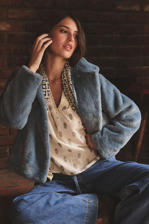 A woman sits in front of a brick wall, touching her hair and looking to the side while wearing the ANA TOP by Velvet by Graham & Spencer, paired with a blue faux fur jacket and wide-leg jeans.