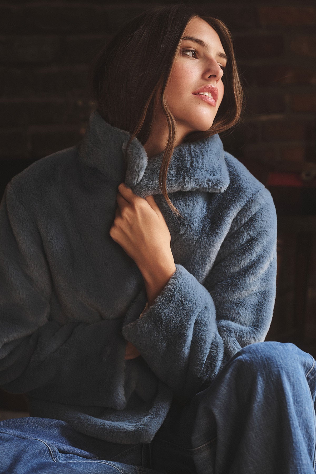 A person wears the SHEENA LUXE FAUX FUR JACKET by Velvet by Graham & Spencer with jeans, sitting indoors and looking to the side while holding the collar for a cozy fit.-39840062046401