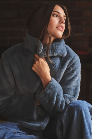 A person wears the SHEENA LUXE FAUX FUR JACKET by Velvet by Graham & Spencer with jeans, sitting indoors and looking to the side while holding the collar for a cozy fit.
