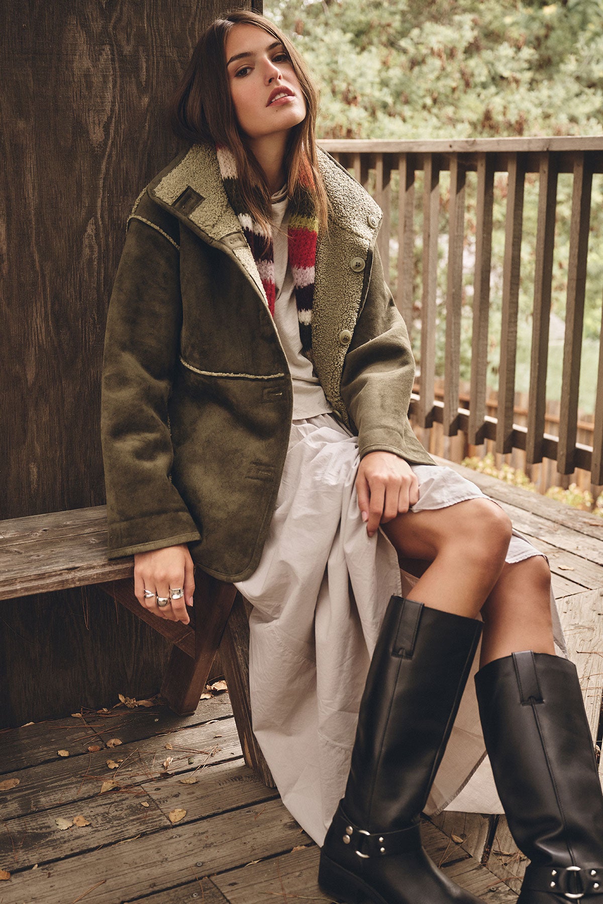 A woman sits on a bench wearing the SHONA SHERPA REVERSIBLE JACKET by Velvet by Graham & Spencer, paired with a long white skirt, striped scarf, and black knee-high boots. Trees and a wooden railing are behind her.-39840000671937
