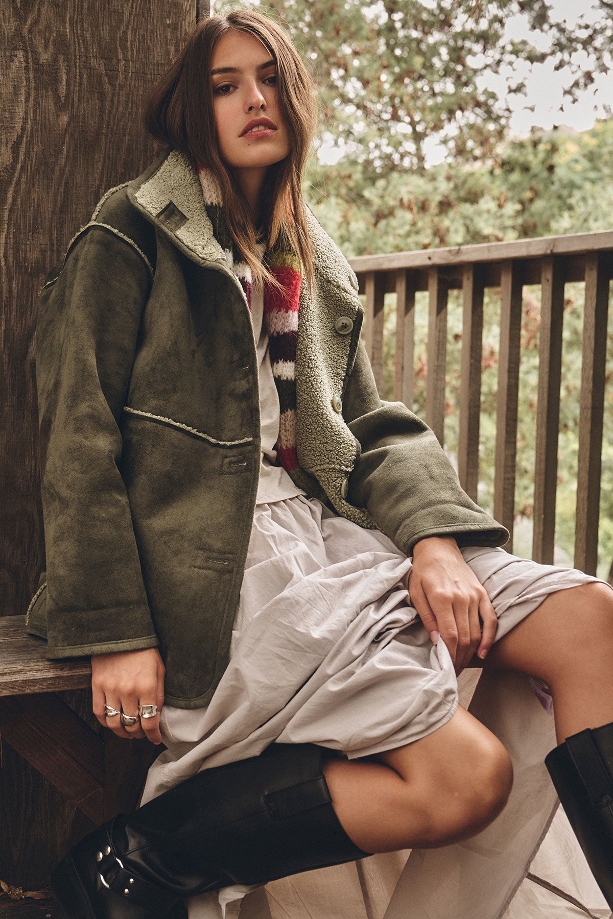  A woman sits on a wooden bench wearing the SHONA SHERPA REVERSIBLE JACKET by Velvet by Graham & Spencer, paired with a striped scarf, light skirt, and black boots. Trees and a wooden railing are in the background. 