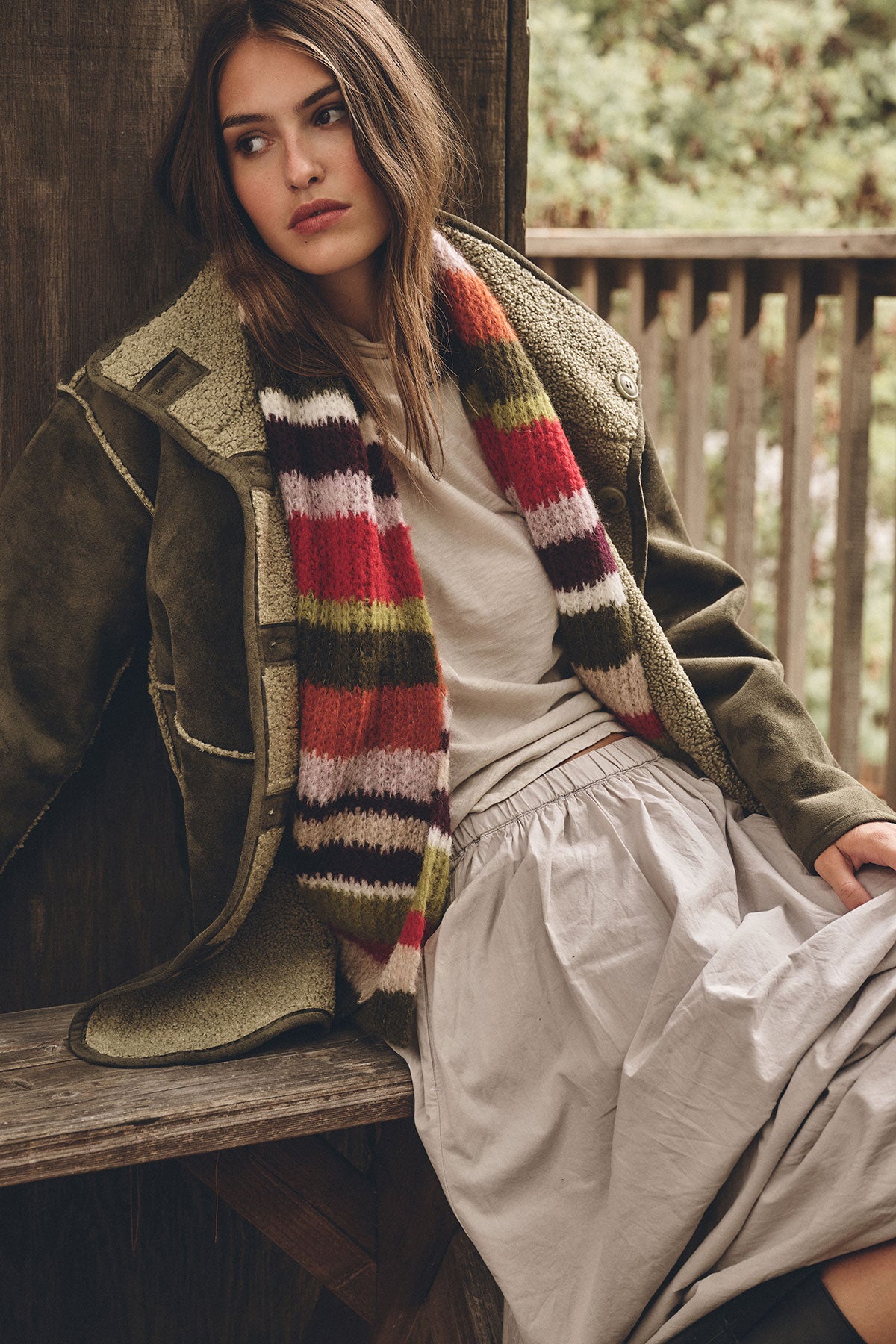 A woman sits on a wooden bench wearing the SAVI TEE by Velvet by Graham & Spencer, a shearling jacket, striped scarf, and light gray skirt with greenery in the background.-39840046678209