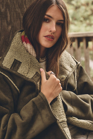 A woman sits outdoors holding the collar of the SHONA SHERPA REVERSIBLE JACKET by Velvet by Graham & Spencer, with a wooden wall and greenery in the background.