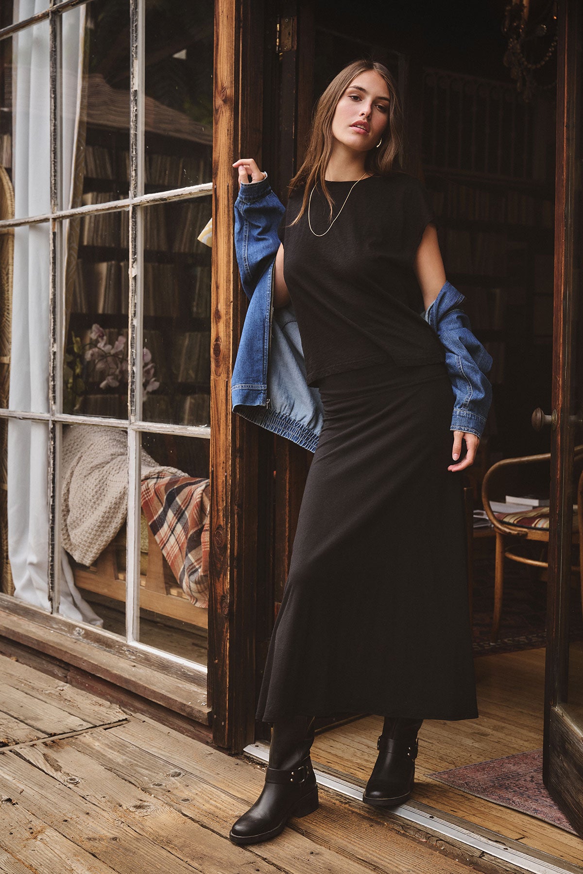 A woman stands in a doorway wearing the LUELLA SKIRT by Velvet by Graham & Spencer, styled with black boots and a blue denim jacket draped over her arms. She poses with one hand on the doorframe, looking at the camera.-39840118046913