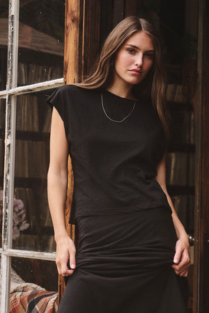 A woman with long brown hair, wearing a black cocoon skirt and the Velvet by Graham & Spencer STERLING TEE, stands in front of a glass door, looking at the camera with a neutral expression.