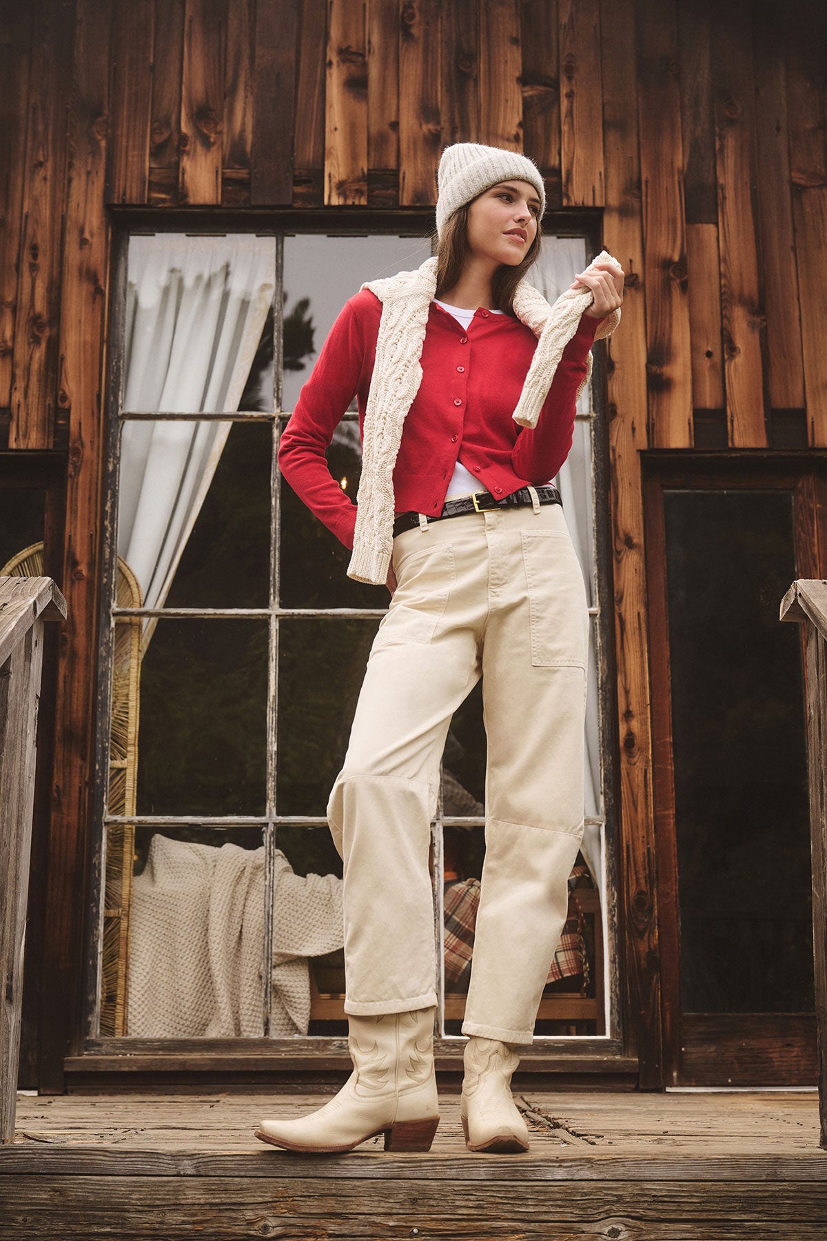 A woman stands on a wooden porch in front of a rustic cabin, wearing the VIOLETTE CARDIGAN by Velvet by Graham & Spencer over cream pants and boots—a perfect choice for lightweight layering.-39839543165121