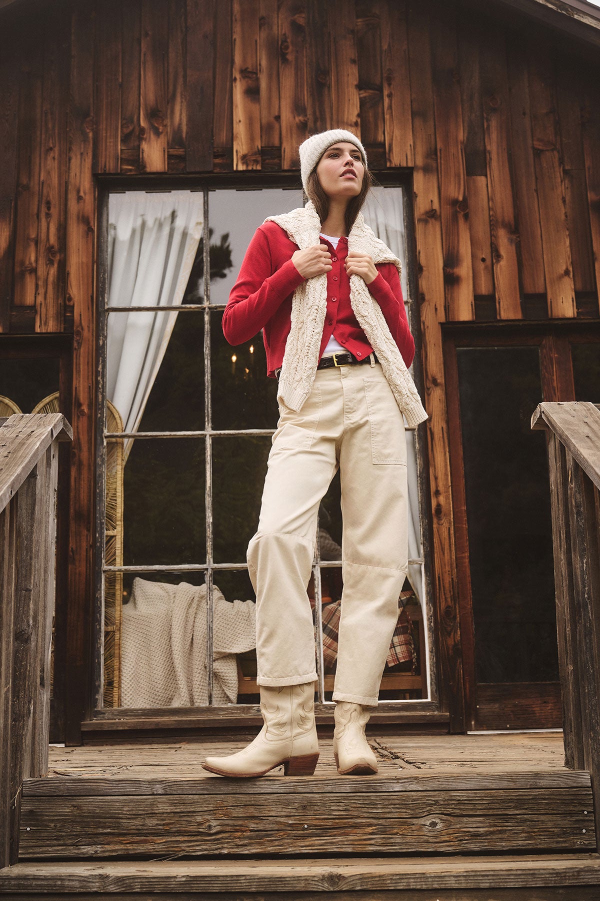 Wearing the KEY BEANIE by Velvet by Graham & Spencer, a person in cream boots, pants, a red shirt, and white vest stands on wooden steps outside a rustic wooden building with large windows.