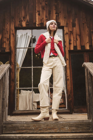 Wearing the KEY BEANIE by Velvet by Graham & Spencer, a person in cream boots, pants, a red shirt, and white vest stands on wooden steps outside a rustic wooden building with large windows.