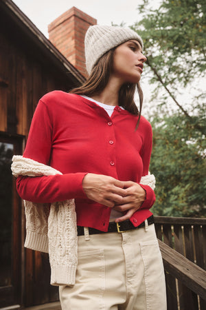 A woman stands outside by a wooden railing wearing the Velvet by Graham & Spencer KEY BEANIE with a red button-up cardigan, white shirt, beige pants, and a sweater draped over her shoulders.