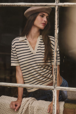 A woman wearing the YAEL Cashmere Sweater Polo by Velvet by Graham & Spencer and a beret leans on a blanket-covered surface, gazing out of a window.