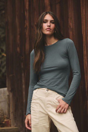 A woman with long brown hair wears the ZOFINA TEE by Velvet by Graham & Spencer and beige pants, standing in front of a wooden wall outdoors.