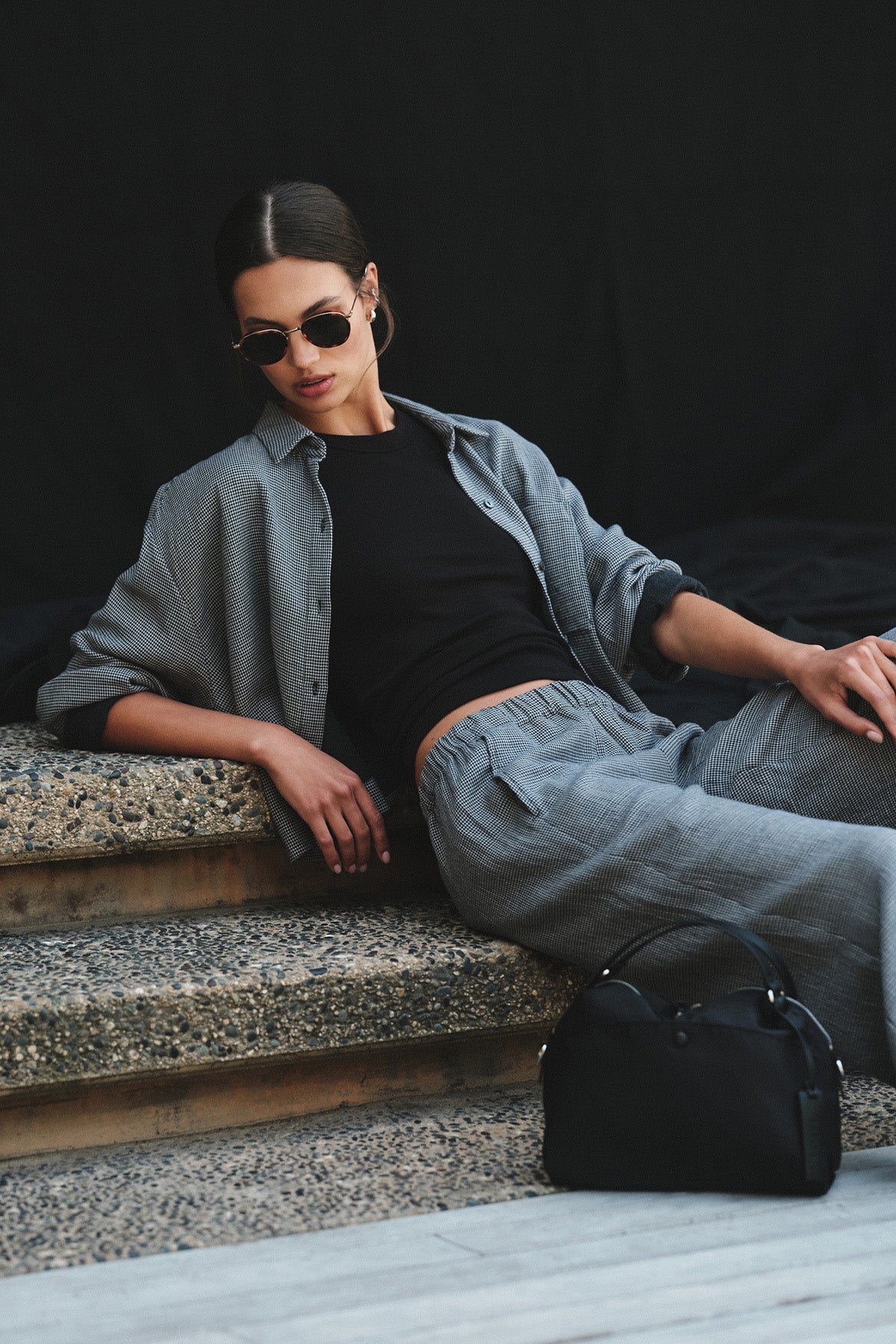 A woman in sunglasses, a black top, and the ADELINE SHIRT with matching pants by Velvet by Graham & Spencer sits on outdoor steps beside a black handbag against a dark background.