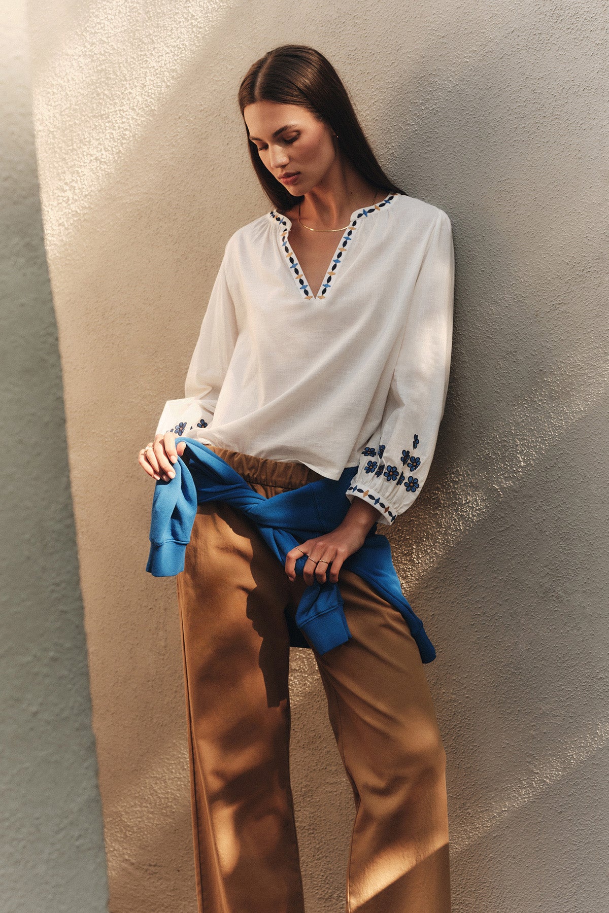 A woman stands against a beige textured wall, wearing the AMANI BLOUSE by Velvet by Graham & Spencer, tan wide-leg pants, and a blue sweater tied around her waist.