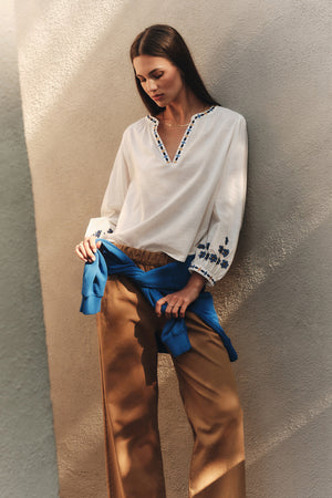 A woman stands against a beige textured wall, wearing the AMANI BLOUSE by Velvet by Graham & Spencer, tan wide-leg pants, and a blue sweater tied around her waist.