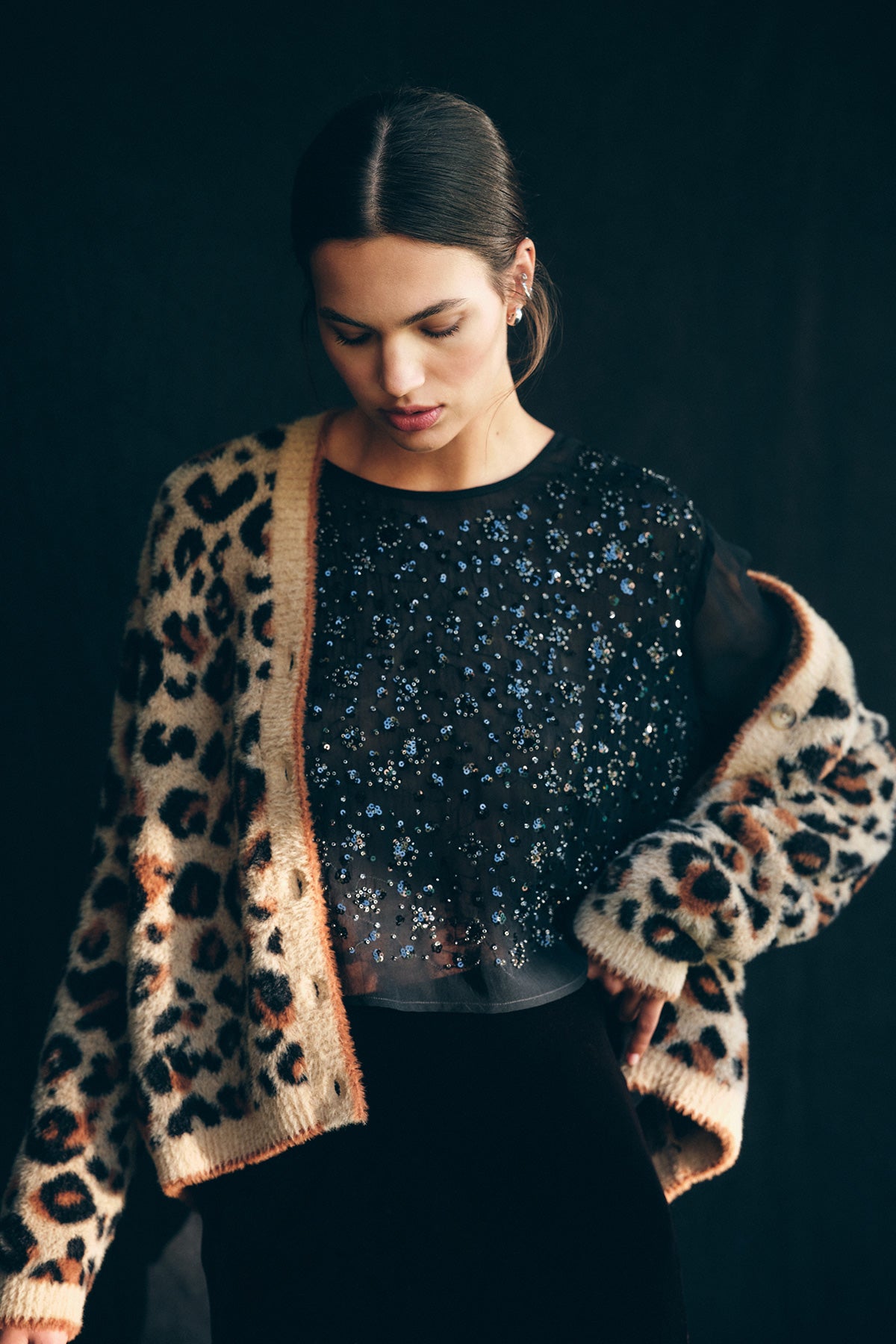 A woman wearing the BETSY CARDIGAN in leopard print by Velvet by Graham & Spencer over a sheer black beaded top, looking down against a dark background.