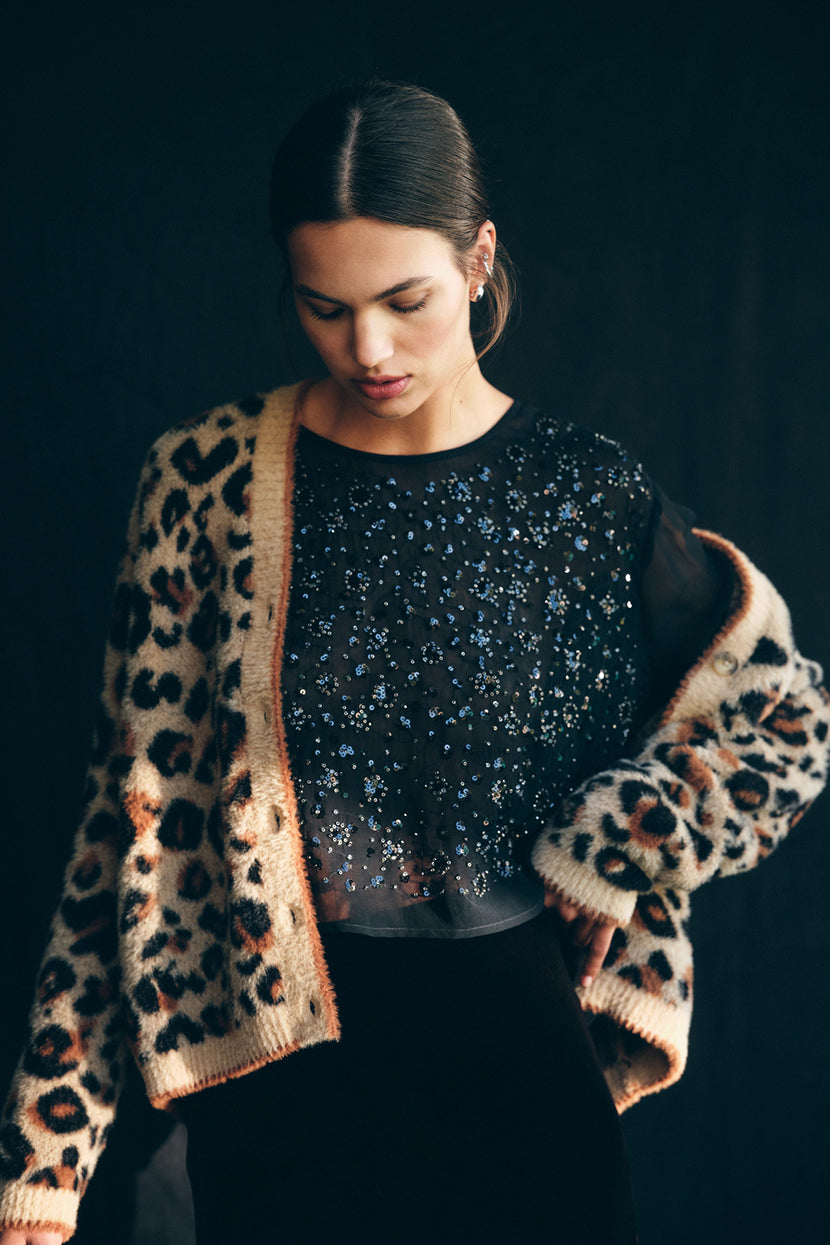 A woman wearing the BETSY CARDIGAN in leopard print by Velvet by Graham & Spencer over a sheer black beaded top, looking down against a dark background.