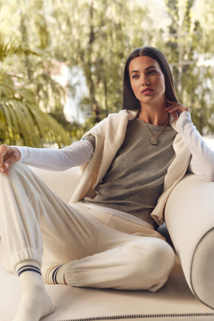 A woman sits on a white sofa outdoors, wearing the BRONSON TEE by Velvet by Graham & Spencer—a grey crewneck modal rib top—paired with white sweatpants and a sweater draped over her shoulders. Trees are visible in the background.