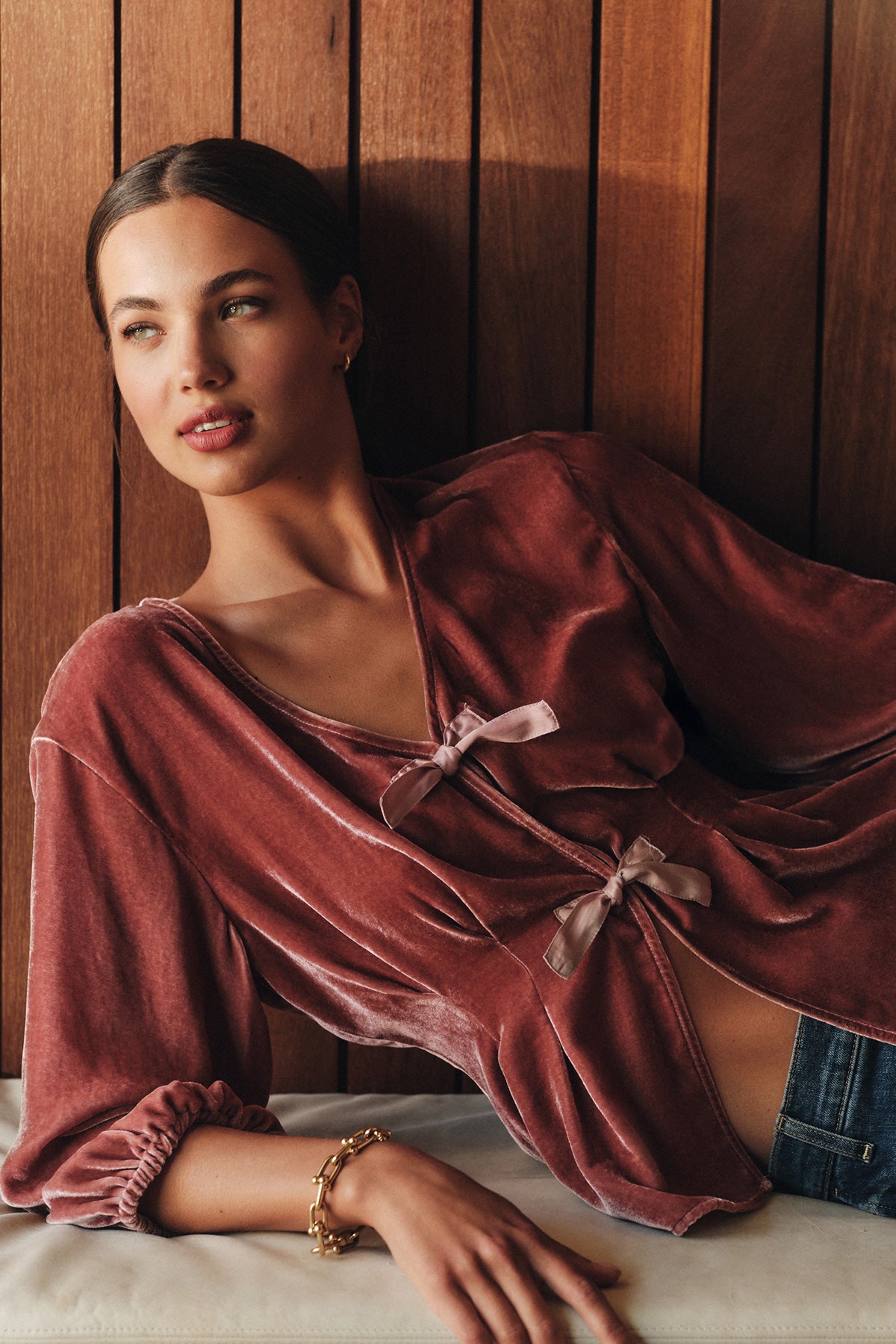 A woman reclines against a wood-paneled wall in the ELIDA TOP by Velvet by Graham & Spencer—its mauve silk velvet and decorative ties paired with jeans and a gold bracelet create a perfect holiday look.