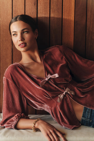 A woman reclines against a wood-paneled wall in the ELIDA TOP by Velvet by Graham & Spencer—its mauve silk velvet and decorative ties paired with jeans and a gold bracelet create a perfect holiday look.