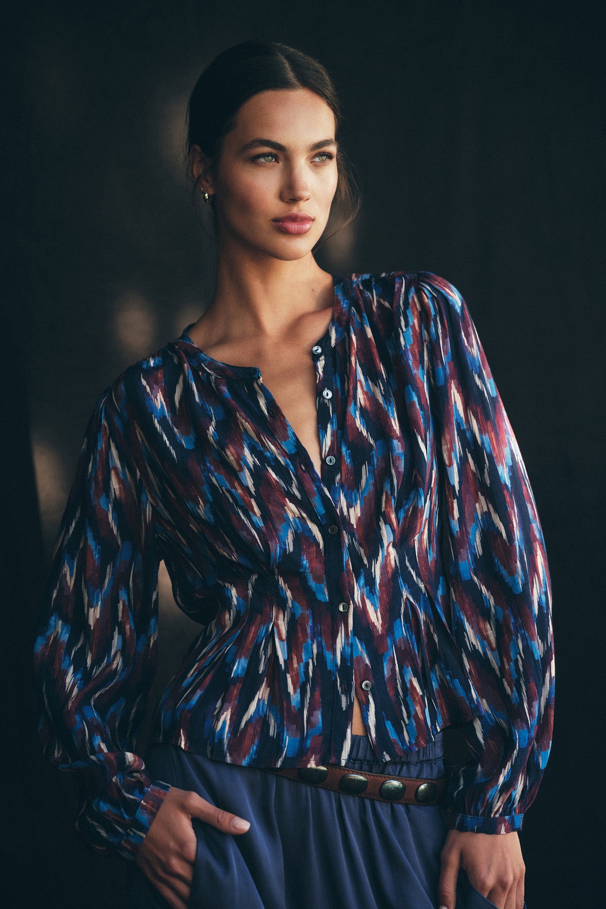 A woman poses against a dark background wearing the ESTELLA BLOUSE from Velvet by Graham & Spencer, a long-sleeve, button-up lightweight cotton blouse featuring a blue, red, white, and black abstract pattern and a belted blue skirt.