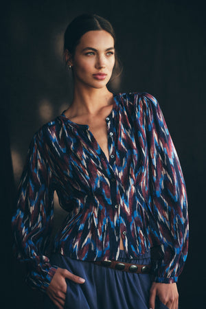 A woman poses against a dark background wearing the ESTELLA BLOUSE from Velvet by Graham & Spencer, a long-sleeve, button-up lightweight cotton blouse featuring a blue, red, white, and black abstract pattern and a belted blue skirt.