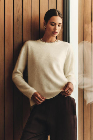 A person wearing the JACQUELINE CASHMERE SWEATER by Velvet by Graham & Spencer, paired with dark pants and gold jewelry, stands indoors beside a wood-paneled wall near a window.
