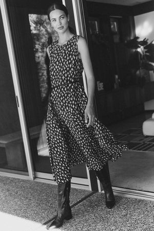 A woman in the KAITLYN DRESS by Velvet by Graham & Spencer, featuring patterned silk cotton voile, stands in tall boots before a sliding glass door, gazing slightly to the side.