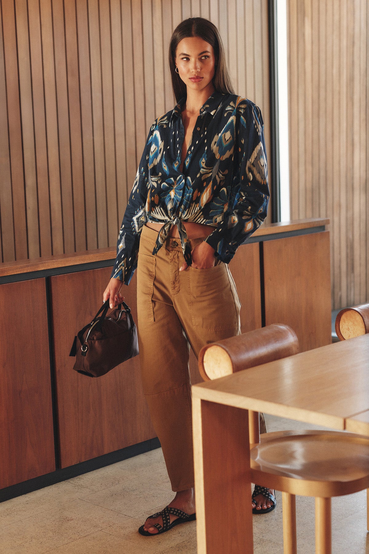 A woman stands indoors wearing the KAYLA TOP by Velvet by Graham & Spencer, a patterned blue button-down blouse, paired with tan high-waisted pants, black sandals, and holding a dark handbag beside a wooden table and chairs.
