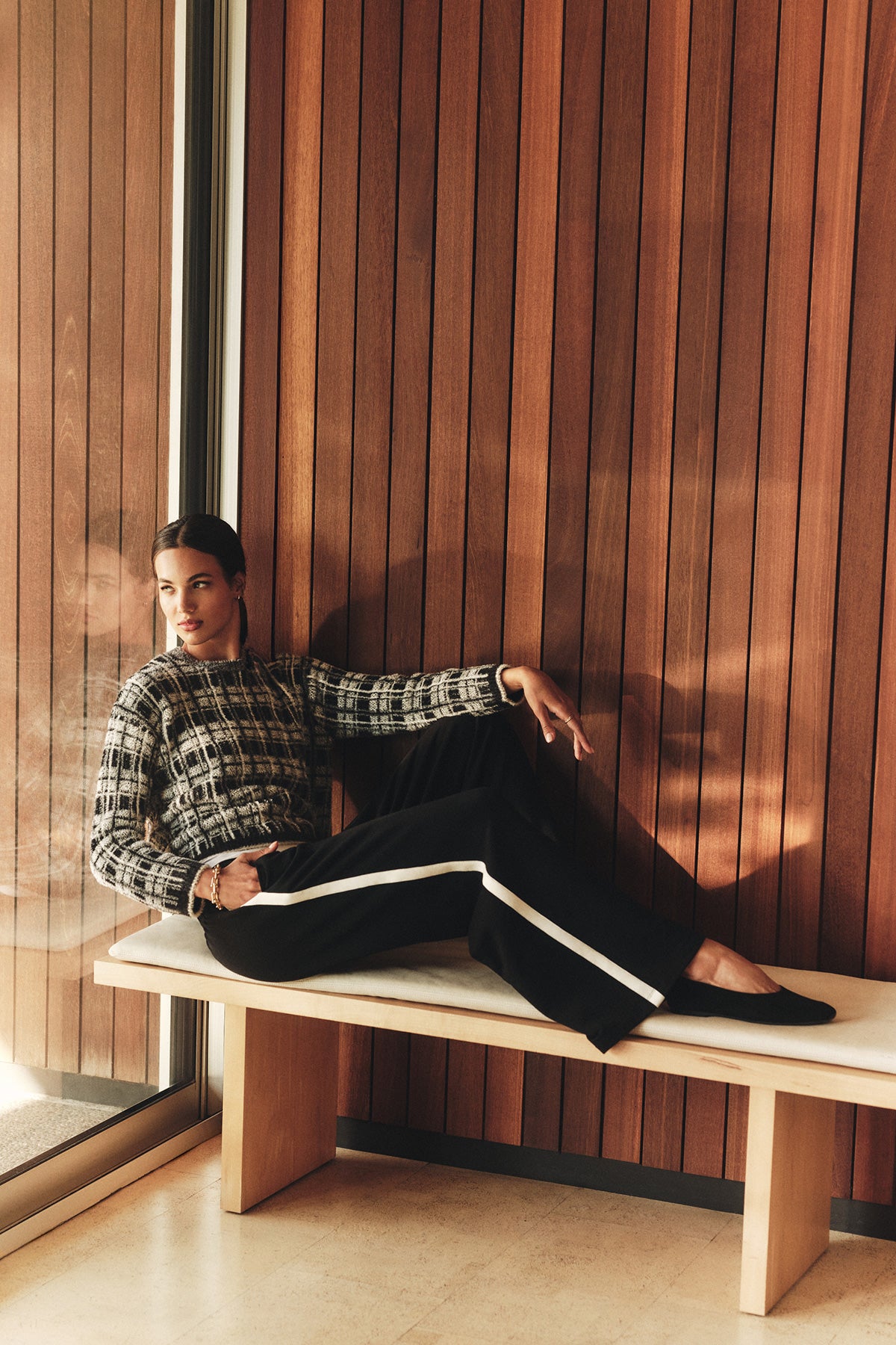 A woman sits on a bench by a wooden wall, wearing the LACEY SWEATER by Velvet by Graham & Spencer with a plaid design, paired with black pants featuring white stripes, looking to the side.-40278603268289