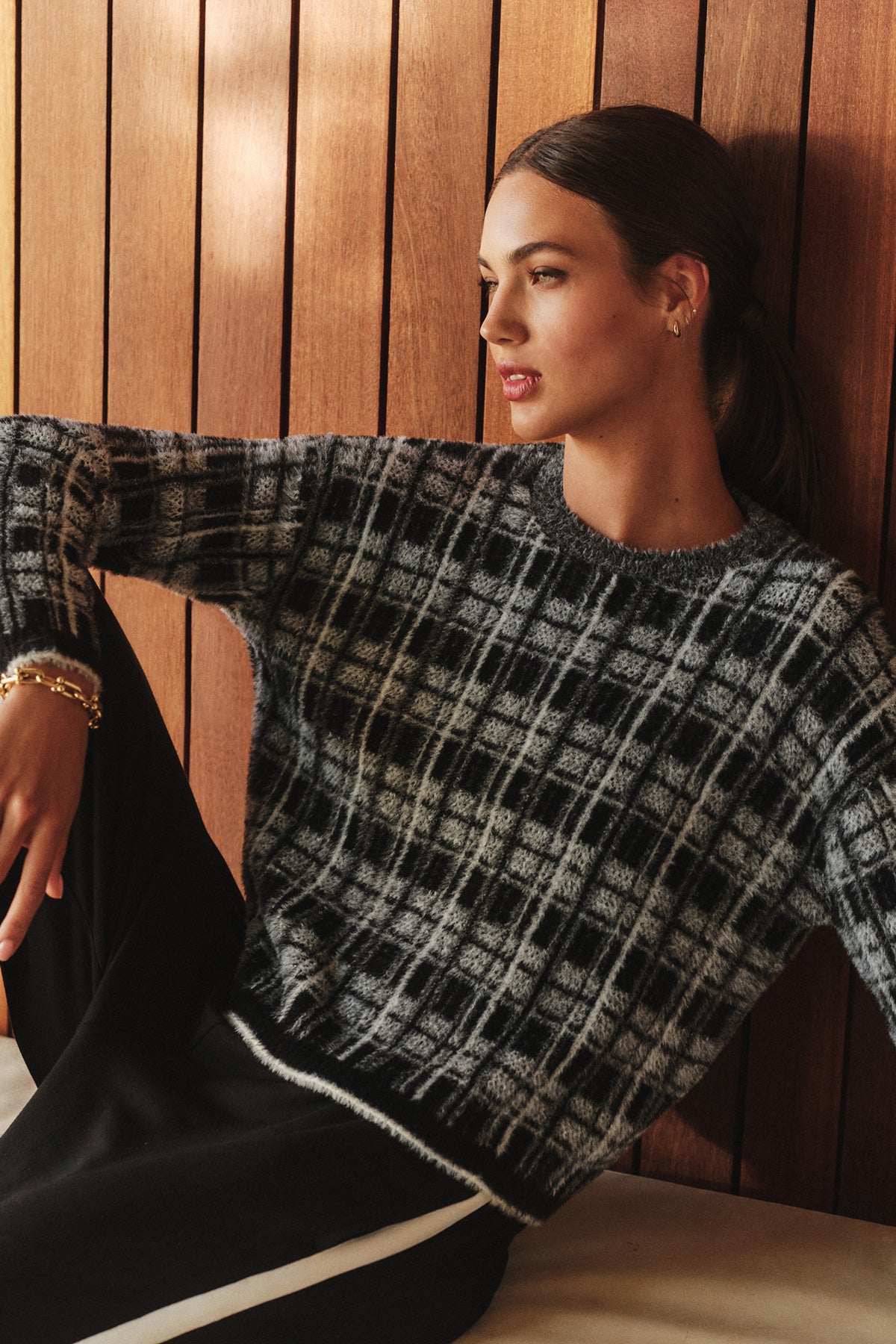 A woman sits against a wooden wall, looking to the side, dressed in the LACEY SWEATER by Velvet by Graham & Spencer—a stylish black and white textured piece—paired with black pants featuring a white stripe.-40278600450241