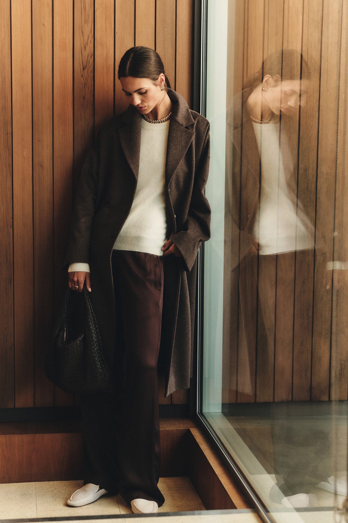 A woman stands indoors by a wood-paneled wall, holding a black textured handbag and wearing the LAFAYETTE COAT by Velvet by Graham & Spencer over a white sweater, wide-leg dark pants, and white shoes.-40263652081857