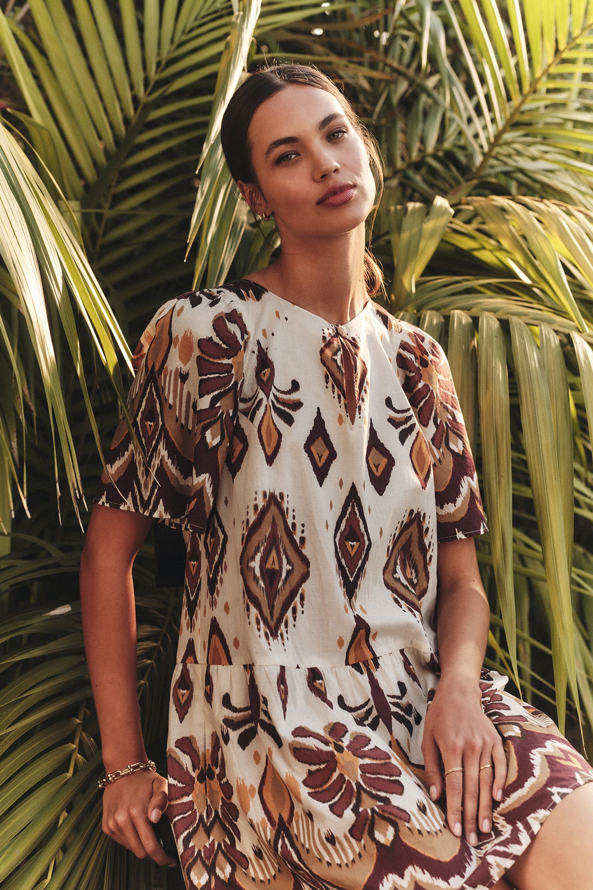 A woman wearing the MAGGIE DRESS by Velvet by Graham & Spencer, featuring a patterned fabric and adjustable belt, sits before large palm leaves and looks at the camera with a neutral expression.-40598485434561