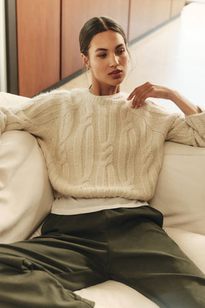 A woman in the MARINA SWEATER by Velvet by Graham & Spencer—cream cable knit with subtle metallic threads—and olive green pants sits on a white couch, looking to the side with one arm resting on the back.