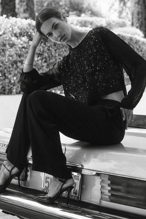 A woman in black pants and the Velvet by Graham & Spencer MARLOWE TOP sits on the trunk of a vintage car outdoors, resting her head on her hand.