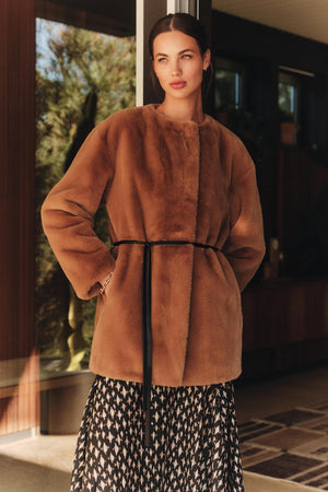 A woman stands indoors wearing the Velvet by Graham & Spencer MARMONT LUXE FAUX FUR JACKET, featuring snap button closures and a thin black belt, paired with a black-and-white patterned skirt.