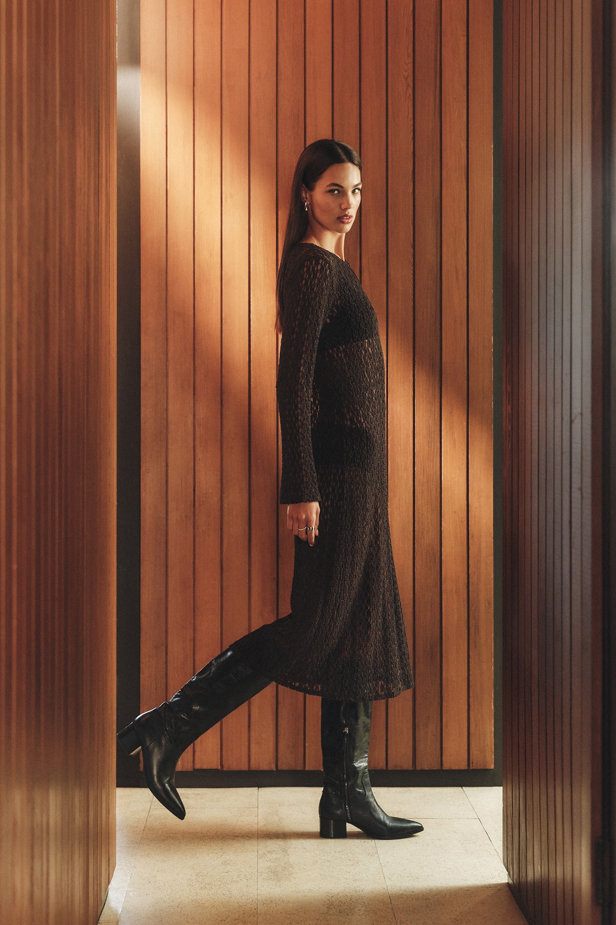 Woman wearing the TABITHA DRESS by Velvet by Graham & Spencer and knee-high black boots stands sideways in a narrow hallway with wooden paneled walls, facing the camera.-40278635020481