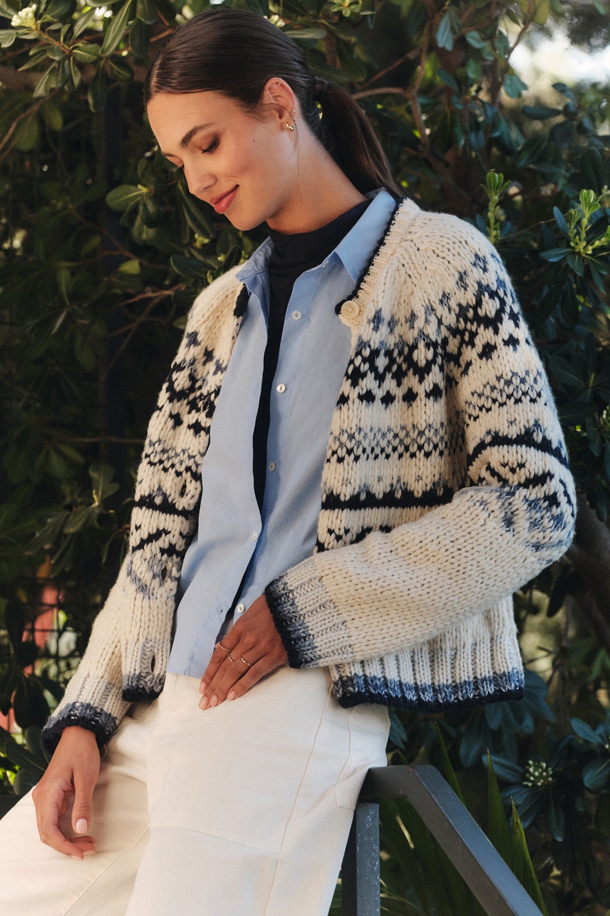 Wearing the TOVAH CARDIGAN by Velvet by Graham & Spencer over a light blue button-up and white pants, a woman stands outdoors near greenery, looking down with a slight smile.-40333273923777