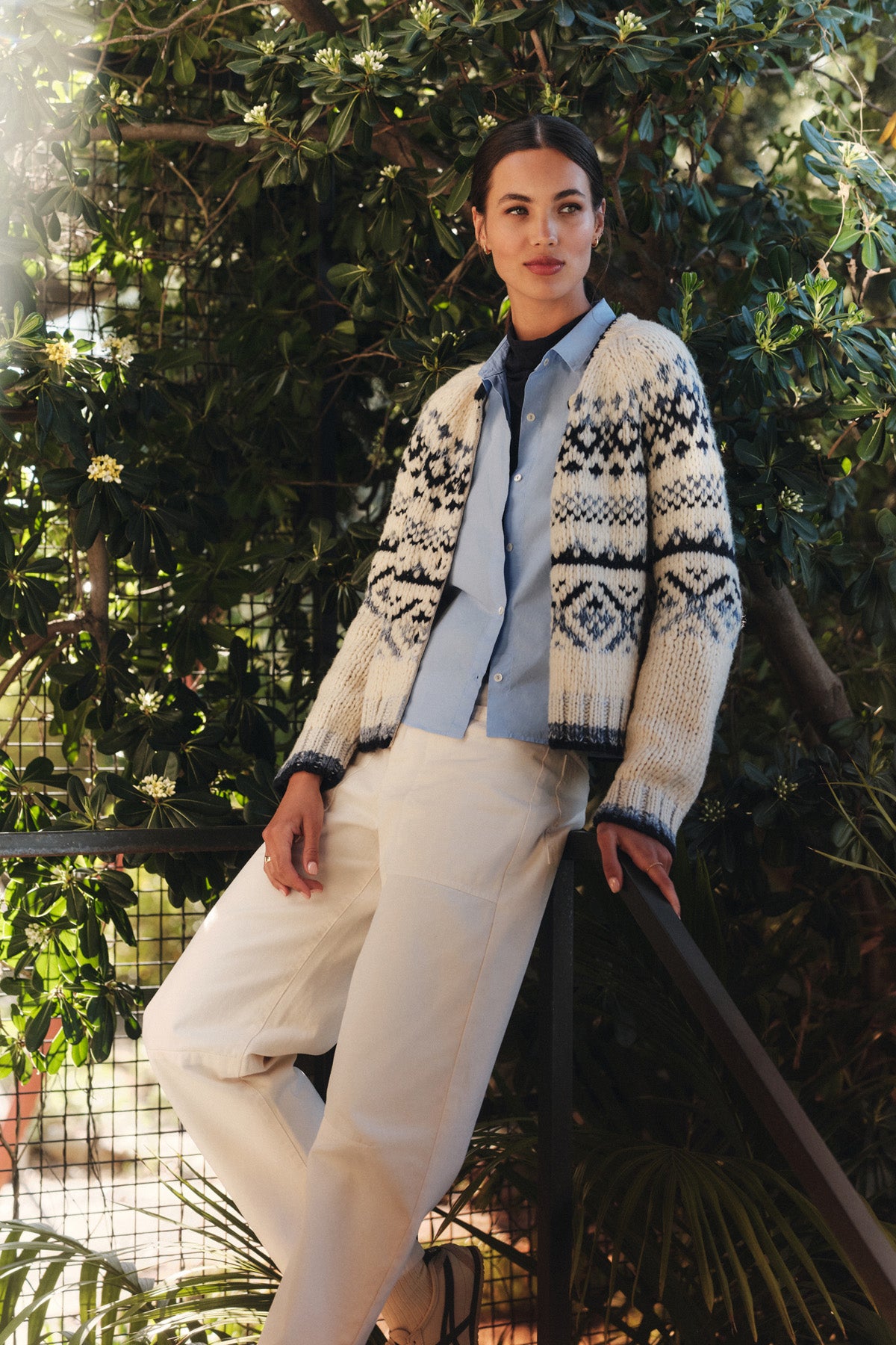 Wearing the TOVAH CARDIGAN by Velvet by Graham & Spencer over a light blue shirt and white pants, a person leans against a railing with greenery in the background.