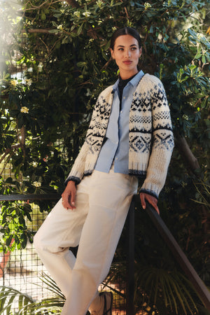 Wearing the TOVAH CARDIGAN by Velvet by Graham & Spencer over a light blue shirt and white pants, a person leans against a railing with greenery in the background.